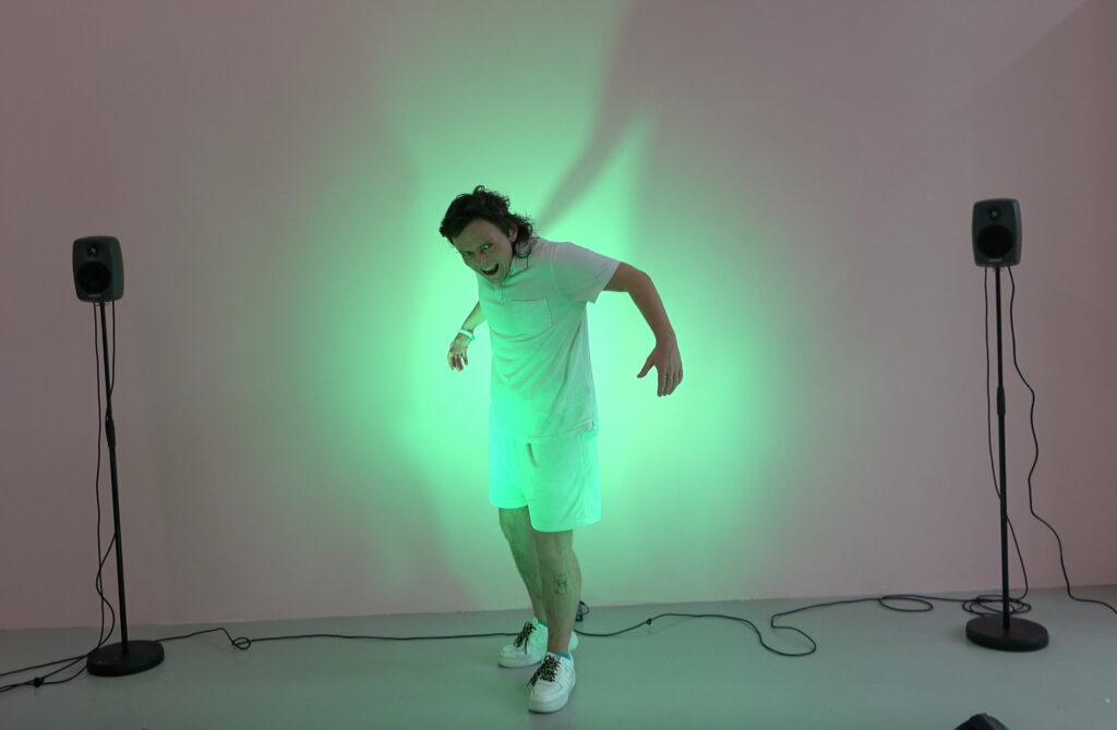A handsome trans-masculine hunk is captured mid performance. They stare menacingly towards you, arms back as though caught mid-flap. They are illuminated by a green light.